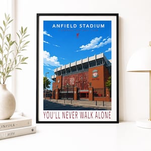 Può includere: Una stampa artistica incorniciata dell'Anfield Stadium, casa del Liverpool Football Club. L'immagine mostra l'esterno dello stadio in mattoni rossi sotto un cielo azzurro con nuvole, lo stemma del Liverpool e il testo "ANFIELD STADIUM" e "YOU'LL NEVER WALK ALONE."