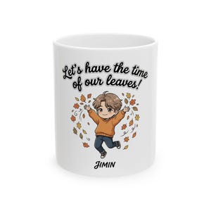 May include: A white ceramic mug featuring a cartoon illustration of a person with brown hair wearing an orange sweater and blue jeans, surrounded by falling autumn leaves. The text reads Let's have the time of our leaves! and JIMIN.