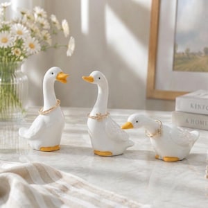 May include: Three white ceramic duck figurines with yellow beaks and feet. Each duck is wearing a gold-toned ring around its neck. The ducks are positioned in a row, with two standing upright and one leaning forward.