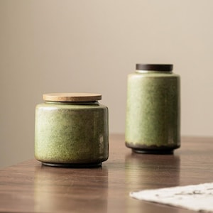 May include: Two ceramic canisters with a mottled light green glaze and dark brown bases. The shorter, wider canister has a flat, light-colored wooden lid, while the taller, narrower canister features a dark brown lid.