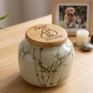 Peut inclure: Une urne pour animal en céramique avec une base crème ornée d'un motif de feuilles de bambou vertes. Le couvercle rond en bois est gravé du nom Zeus, d'une illustration au trait d'une silhouette de chien et de chat, et des années 2010-2024.