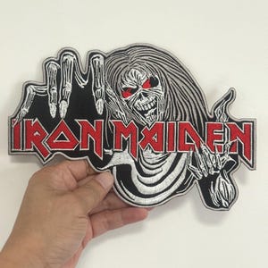 May include: An embroidered patch featuring the Iron Maiden logo in red with a silver border. The design includes a skeletal figure with long hair, red eyes, and clawed hands reaching out, all rendered in black, white, and silver thread on a black background.