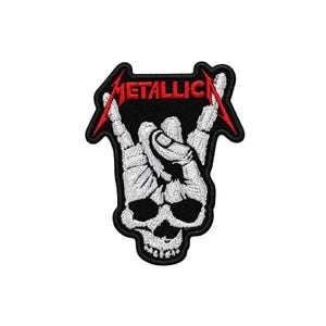 Può includere: Una toppa ricamata raffigurante una mano scheletrica bianca che fa un segno rock and roll emergendo da un teschio. La parola METALLICA è ricamata in filo rosso nella parte superiore su uno sfondo nero con un bordo nero spesso.