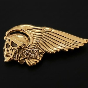 Könnte beinhalten: Eine goldfarbene Metallbrosche, die einen Totenkopf mit geflügeltem Helm darstellt. Das Design enthält ein Banner im unteren Bereich mit dem geprägten Text HELLS ANGELS. Das Objekt hat eine polierte, reflektierende Oberfläche und ein detailliertes Design.