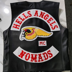 Op de afbeelding: Een zwart leren vest met geborduurde patches op de achterkant. De bovenste patch bevat de tekst HELLS ANGELS in rode letters op een witte achtergrond. Daaronder bevindt zich een gevleugeld schedel-embleem in geel, rood en wit met een MC-patch.