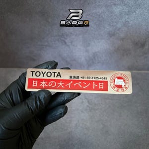 May include: A rectangular metal badge with a brushed finish featuring the text TOYOTA, a phone number, and Japanese characters on a red background. A circular logo with Japanese text is on the right side, and the BADGR logo is at the top.