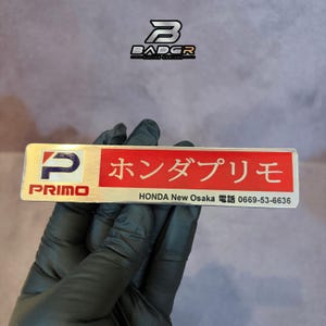 May include: A rectangular metallic emblem featuring a blue and red logo on the left with the text PRIMO below it. The right side has a red background with white Japanese text, followed by the black text HONDA New Osaka and phone number 0669-53-6636.
