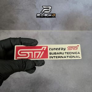 May include: A rectangular emblem featuring a red STI logo on the left and the text tuned by STI SUBARU TECNICA INTERNATIONAL in black on a cream background. The emblem has a clean, flat design with bold lettering and a distinct automotive aesthetic.