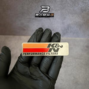 May include: A rectangular metal emblem featuring the K&N logo with a horizontal orange stripe above a red stripe. The text PERFORMANCE FILTERS is printed in black below the stripes. The emblem has a brushed silver finish and rounded corners.