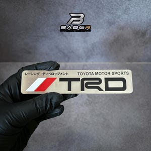 Op de afbeelding: Een rechthoekig metalen embleem met de zwarte tekst TRD en TOYOTA MOTOR SPORTS. Links van de tekst staat een grafische afbeelding met een witte en rode diagonale streep. Boven de tekst staan Japanse karakters: レーシング・ディベロップメント.