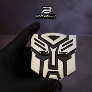 Könnte beinhalten: Ein metallisches silber-schwarzes Emblem mit einem stilisierten Robotergesicht-Design und scharfen geometrischen Linien. Das Emblem hat eine polierte Oberfläche und wird mit dem Text BADGR CUSTOM EMBLEMS oben im Bild angezeigt.