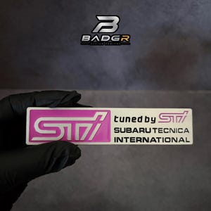 Pode incluir: Um emblema retangular com um logótipo STI rosa e branco no lado esquerdo. O lado direito apresenta o texto tuned by STI, SUBARU TECNICA INTERNATIONAL em letras pretas. O emblema tem um design plano e limpo com bordos definidos.