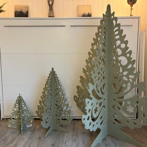 Könnte beinhalten: Drei freistehende dekorative Bäume aus hellgrünem Holz mit komplizierten lasergeschnittenen Mustern. Die Bäume variieren in der Höhe, wobei der kleinste Baum kleine integrierte warmweiße LED-Lichter entlang seiner zentralen Struktur aufweist.