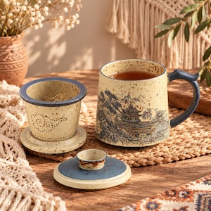 May include: A ceramic tea mug with a speckled cream finish, featuring a blue handle and a detailed dark blue illustration of a traditional pagoda building. The set includes a matching removable tea infuser basket and a circular lid with a blue rim.