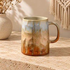 Puede incluir: Una taza de cerámica con superficie texturizada y facetada que presenta un esmalte reactivo que transiciona de un azul pálido en el borde a un tono bronceado cálido en el centro y un naranja óxido profundo en la base, con asa curva.
