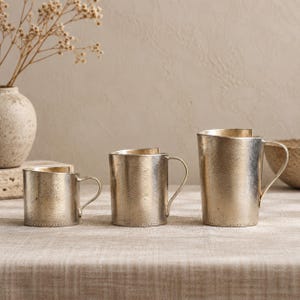 May include: Three metallic mugs with a textured, weathered gold and silver finish. Each mug features a cylindrical body, a curved handle, and a slightly uneven top rim. The mugs are arranged in a row, varying in height from shortest to tallest.