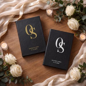 Puede incluir: Dos libros de votos rectangulares negros con cubiertas texturizadas. El libro de la izquierda presenta un monograma dorado OS con el texto SOPHIA'S VOWS 11.11.2026. El libro de la derecha presenta un monograma blanco OS con el texto OLIVER'S VOWS 11.11.2026.