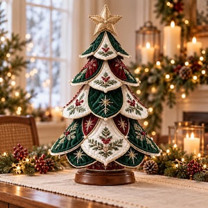 Peut inclure: Un sapin de Noël décoratif de table composé de couches de tissu matelassé vert foncé, bordeaux et crème. L'arbre est orné de broderies dorées complexes, de baies rouges et de petites perles blanches, surmonté d'une étoile en tissu doré sur un socle.
