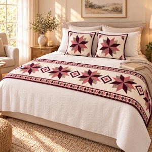 May include: A white bedspread featuring a decorative runner with a repeating star and diamond pattern in shades of burgundy, mauve, and pink. Two matching square throw pillows with the same star design are placed against the headboard.
