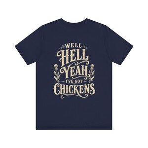 Op de afbeelding: Een marineblauw t-shirt met korte mouwen en een crèmekleurige grafische print op de borst. Het ontwerp bevat de tekst WELL HELL YEAH I'VE GOT CHICKENS in een decoratief vintage lettertype, geflankeerd door twee geïllustreerde distelplanten.