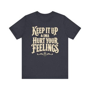 May include: A dark heather gray short-sleeve t-shirt featuring the cream-colored text "KEEP IT UP & IMA HURT YOUR FEELINGS" in a bold, stylized font, centered above a small decorative thistle graphic with symmetrical flourishes on the chest.