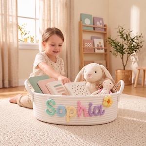May include: A white woven rope storage basket featuring the name Sophia in colorful yarn lettering. The basket is filled with several books, a plush rabbit toy, and a small doll. A yellow fabric flower is attached to the side of the basket.