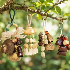 May include: Four hanging wind chimes suspended from a tree branch. Each features a gold metal ring, natural fibre loops, and a combination of brown coconut shells, wooden beads, small metal bells, and cream-coloured fabric tassels.