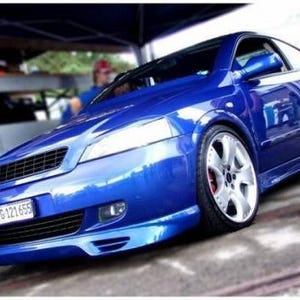 Pode incluir: Um carro coupé azul brilhante com um kit de carroçaria rebaixado, jantes de liga leve prateadas de vários raios e pinças de travão vermelhas. A matrícula dianteira apresenta o texto TG-121655. Grelha frontal preta e faróis transparentes.