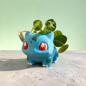 May include: A light blue creature-shaped planter with large red eyes and dark green spots on its back and legs. The planter features a wide opening on its back containing a small green plant with round leaves and tiny white flower clusters.