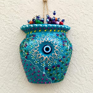 May include: A turquoise ceramic wall planter featuring a central blue and white evil eye motif surrounded by painted floral vines, yellow dots, and pink polka dots. The top is filled with small, colourful glass evil eye beads on wire stems.
