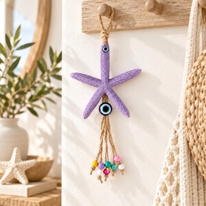 May include: A purple starfish wall hanging featuring a small blue evil eye bead at the top and another evil eye bead suspended below the starfish. The piece includes a jute rope hanger and a tassel of colorful wooden beads in yellow, purple, pink, white, and teal.