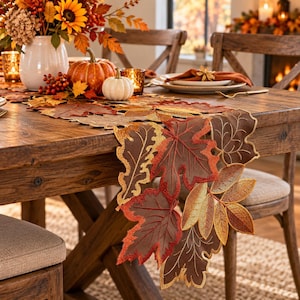 May include: A brown fabric table runner featuring embroidered autumn leaf shapes in shades of orange, red, and gold. The runner is displayed on a wooden table with a white vase, orange and white pumpkins, and place settings with napkins.