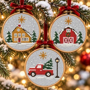 May include: Three circular wooden embroidery hoops containing cross-stitch patterns of a yellow house, a red barn, and a red truck carrying a pine tree. Each hoop features a light grey background, a gold star, and a red velvet ribbon for hanging.