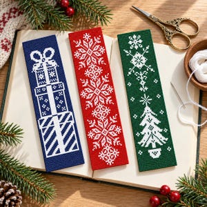 May include: Three rectangular cross-stitch bookmarks featuring white embroidered designs on solid backgrounds. The blue bookmark shows stacked gift boxes, the red one displays a series of snowflakes, and the green one depicts a Christmas tree.