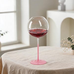 Puede incluir: Una copa de vino de cristal transparente con un cuenco grande y esférico lleno de vino tinto oscuro. El tallo y la base circular son de un color rosa claro opaco y sólido, creando un contraste marcado con la parte superior transparente.