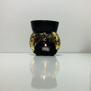May include: A black wax melt warmer with a rounded body and a wide top opening. The exterior features a textured gold leaf finish applied over the black base. A small, lit tea light candle is visible inside the arched opening at the bottom.