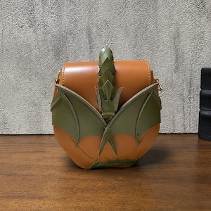 Può includere: Una borsa a tracolla arrotondata color cuoio con inserti in pelle verde oliva a forma di ali di drago sul davanti, un dettaglio centrale verticale simile a una spina dorsale e una piccola coda sul fondo, con attacchi in metallo dorato.