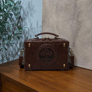 May include: A dark brown rectangular box-shaped bag with a top handle and a detachable shoulder strap. The front features a circular embossed design of a tree surrounded by runes. Four small brass-coloured skull accents adorn the corners.