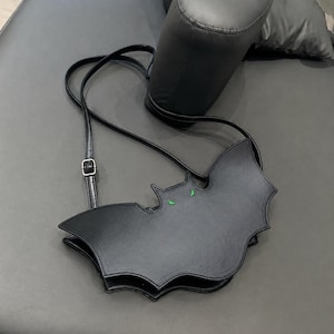 Peut inclure: Un sac bandoulière noir en forme de chauve-souris avec une finition lisse et deux petits yeux verts pointus brodés sur le devant. Le sac comprend une fine bandoulière noire réglable avec une boucle en métal argenté pour ajuster la longueur.