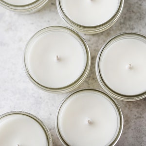 May include: Six clear glass jars with white candles and wicks. The jars are arranged in a circle on a light gray surface.