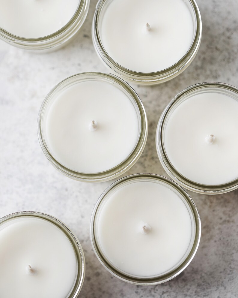 May include: Six clear glass jars with white candles and wicks. The jars are arranged in a circle on a light gray surface.