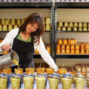 May include: A woman pours wax into ceramic candle jars. The jars are arranged on a table with wicks sticking out. The woman is wearing a white shirt and a black apron. The jars are decorated with a floral pattern.