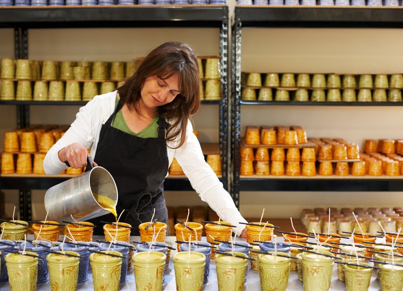 May include: A woman pours wax into ceramic candle jars. The jars are arranged on a table with wicks sticking out. The woman is wearing a white shirt and a black apron. The jars are decorated with a floral pattern.