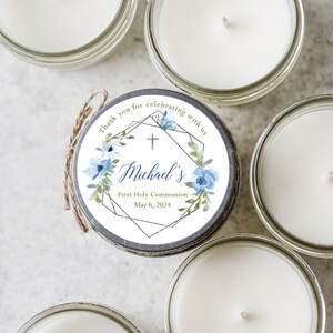 May include: A set of four small glass jars with white candles. Each jar has a round label with a blue and green floral design, a gold geometric frame, and the text "Thank you for celebrating with us + Michael's First Holy Communion May 6, 2024".