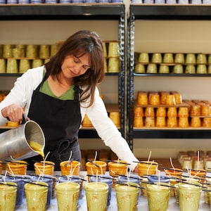 May include: A woman pours wax into ceramic candle jars. The jars are arranged on a table in rows. The jars are decorated with a floral pattern and have a yellow glaze. The woman is wearing a white shirt and a black apron.