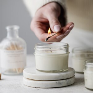May include: A hand lights a white candle with a match. The candle is in a clear glass jar and is sitting on a white marble coaster. There are other candles in the background.