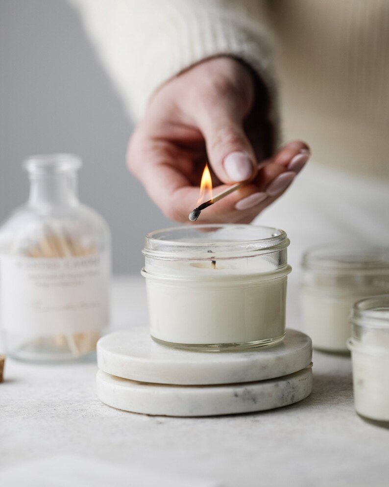 May include: A hand lights a white candle with a match. The candle is in a clear glass jar and is sitting on a white marble coaster. There are other candles in the background.