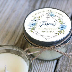 May include: A small glass jar candle with a white label. The label has a geometric design with a cross, blue flowers, and gold accents. The text on the label reads "Thank you for celebrating with us Jason's First Communion May 5, 2019".