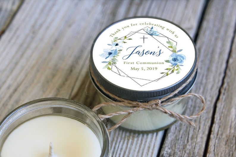 May include: A small glass jar candle with a white label. The label has a geometric design with a cross, blue flowers, and gold accents. The text on the label reads "Thank you for celebrating with us Jason's First Communion May 5, 2019".