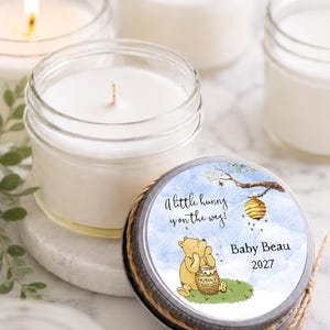 May include: A clear glass jar candle with a white wax fill and a metal lid. The lid features a circular label with a watercolor illustration of a yellow bear, a beehive, and the text A little hunny is on the way! Baby Beau 2027.